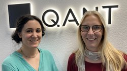 Portrait von Lara Torralbo-Campo und Sandra Mebben vor dem Logo der Firma Q.ANT GmbH