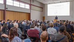 Ein Redner steht auf einer Bühne vor einem großen Publikum im Saal.