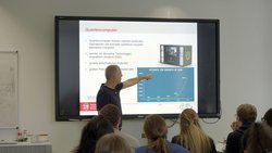 Person vor einem Bildschirm mit Präsentation hält einen Vortrag in einem Seminarraum