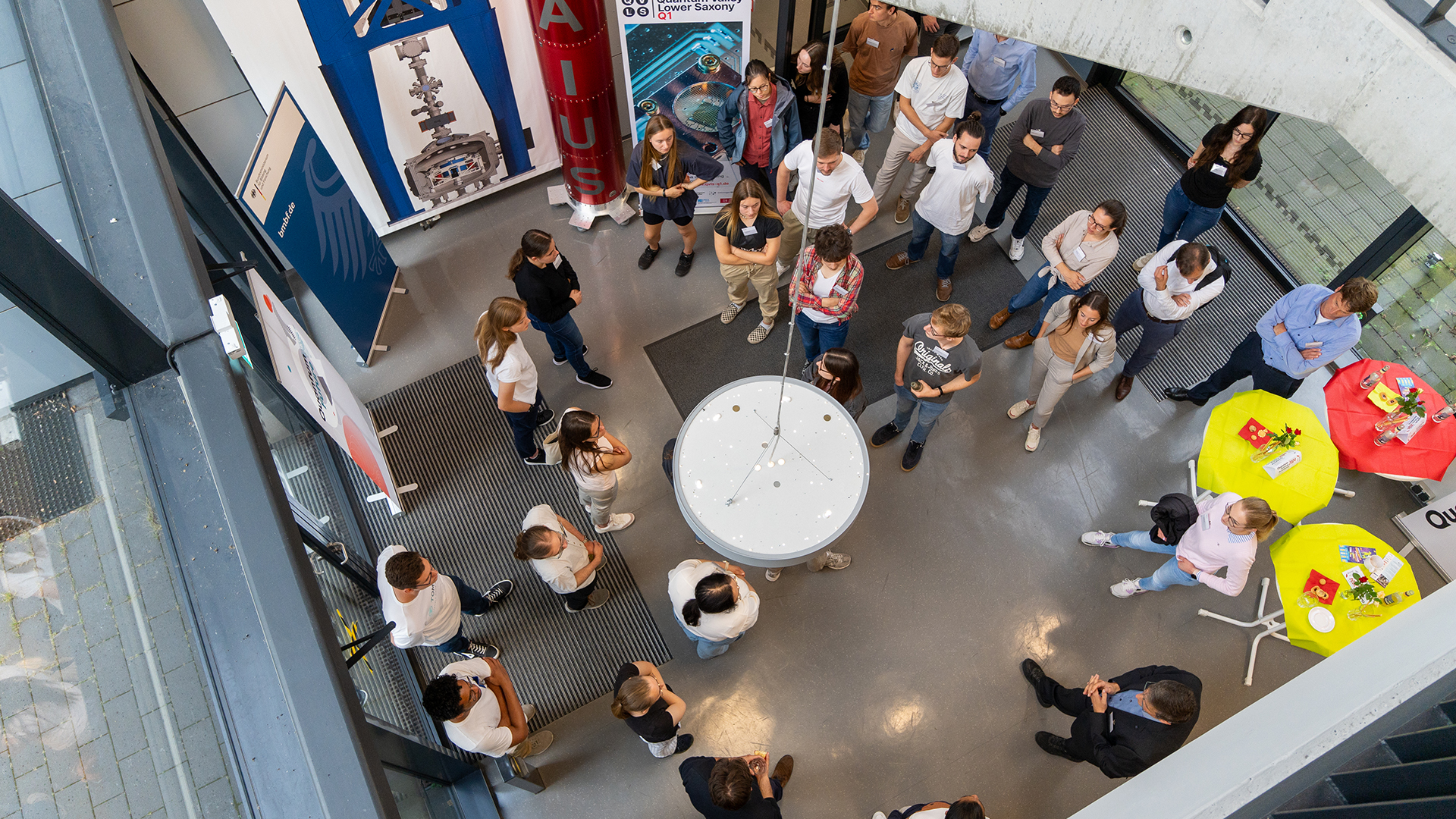 Von oben fotografiert eine Gruppe junger Menschen in einem Foyer. Sie stehen im Kreis und hören einer Person in der Mitte zu.
