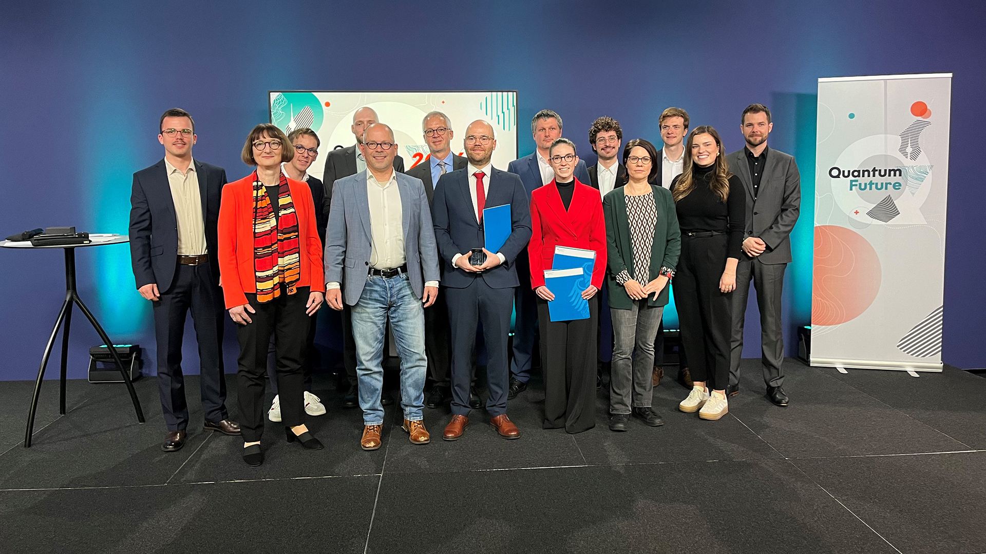 Gruppenfoto: Zehn Personen stehen in zwei Reihen und schauen in die Kamera. Rechts daneben ein Roll-Up mit der Aufschrift "Quantum Future".