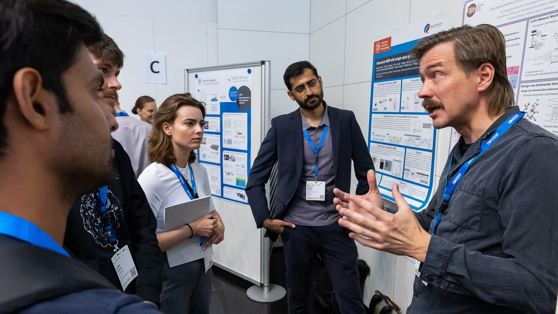 Ein Mann steht vor zwei wisschenschaftlichen Postern. Er erklärt drei jungen Mönnern und einer jungen Frau etwas mit den Händen. Im Hintergrund weitere wissenschaftliche Poster.